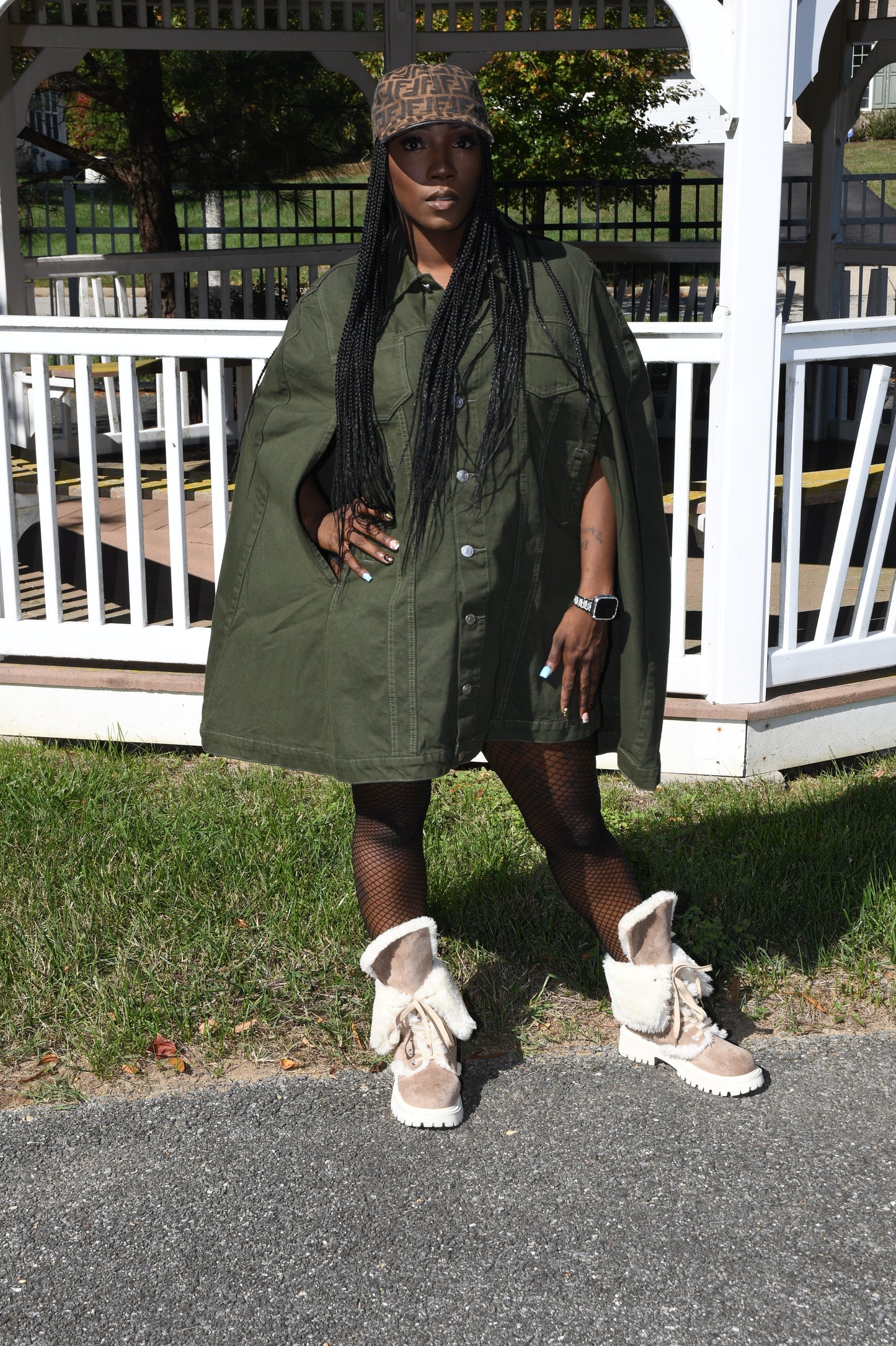 Person wearing a green jacket and white shoes on a street with a white fence and grass in the background.